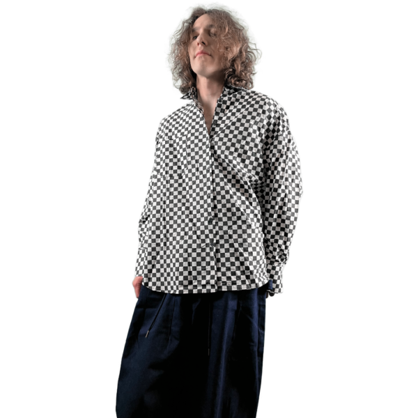 A person with shoulder-length curly hair wearing a black and white checkered shirt looking upwards to the left, standing against a white background.