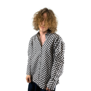 A person with shoulder-length curly hair wearing a black and white checkered shirt standing against a white background, facing slightly to the left with their head tilted downward, obscuring their face.